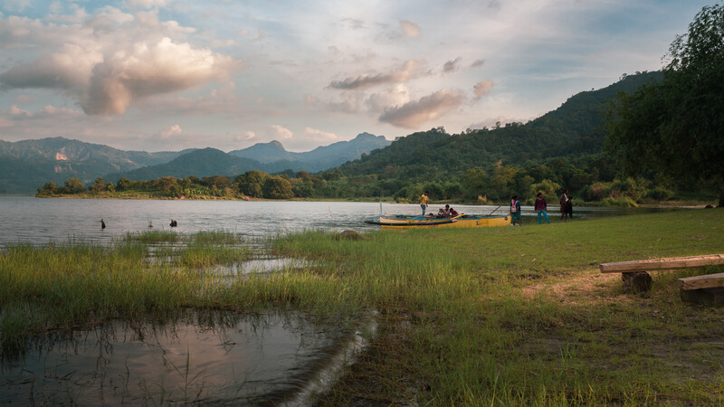 Lakeside landscape with mountains, boats, and green meadow during golden hour with dramatic clouds.
