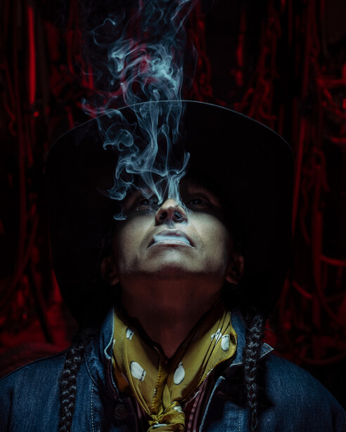 Portrait of person wearing sunglasses with blue smoke flowing upward against dark red background.