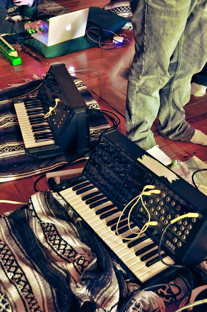 Overhead view of synthesizers and music production equipment arranged on a patterned rug.