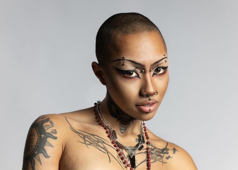 Person with shaved head, dramatic winged eyeliner, multiple tattoos, and beaded necklaces against neutral background.
