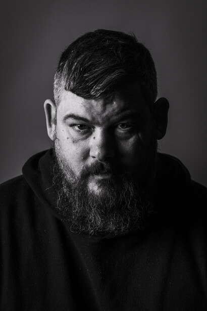 Bearded man with dark hair in black hoodie against dark gray background, looking downward.