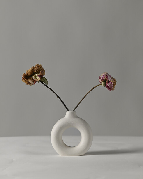 White circular vase with two dried flowers in black stems against neutral background.