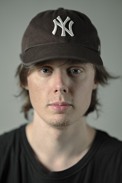 Headshot portrait of a person wearing a dark Yankees cap and black shirt against a neutral gray background.