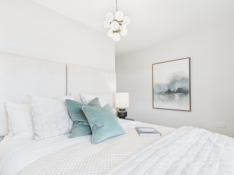 Minimalist bedroom with white bedding, teal throw pillows, pendant light, and framed landscape artwork.