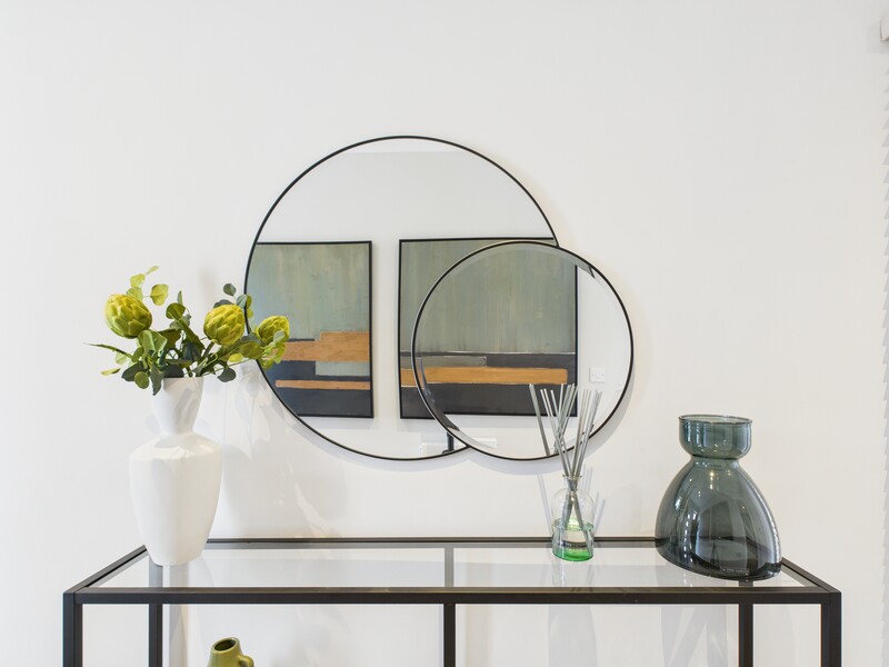 Styled tabletop still life with circular mirrors, white vase, and glass bottles against white wall.