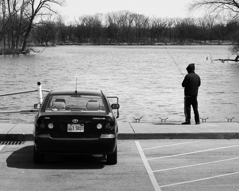 Black and white photo of a man fishing near his parked car