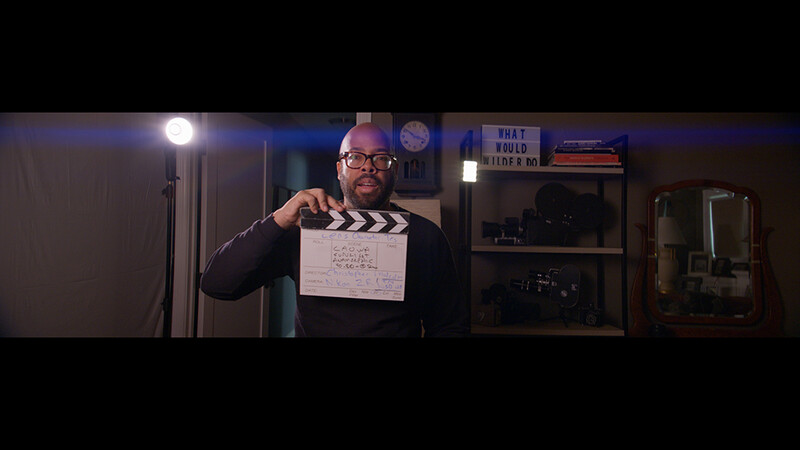 Filmmaker holding a clapperboard in a studio setting with professional lighting and equipment visible in the background.