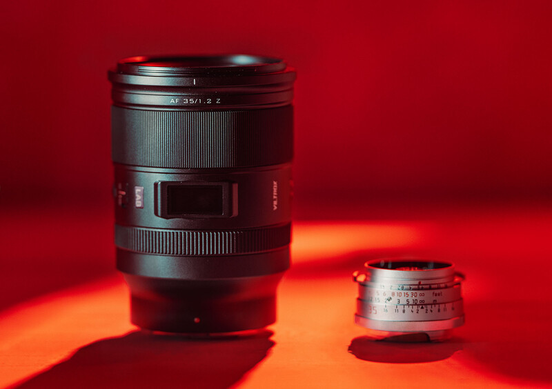 Two camera lenses arranged on a red surface with warm orange directional lighting creating dramatic shadows.
