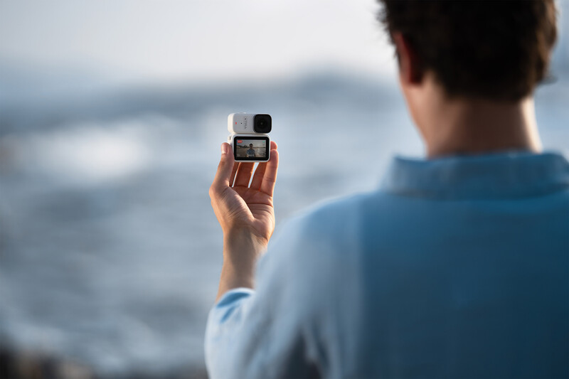 Person holding an action camera aloft while facing a blurred ocean and coastline behind them.