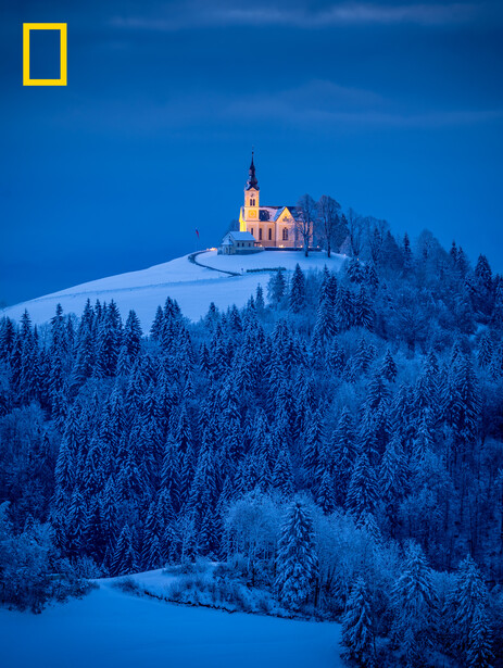 Illuminated church on a snow-covered hill surrounded by dense evergreen forest at twilight.