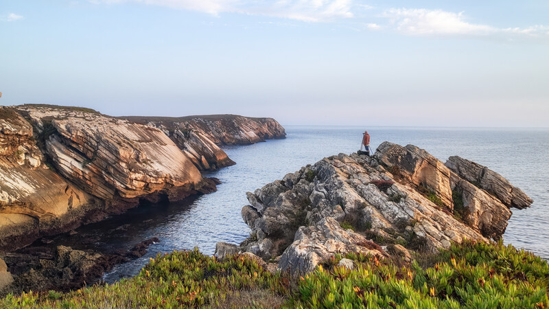 Dramatic rocky coastal cliffs and sea stacks under golden hour light with a lone figure standing on a distant outcrop.
