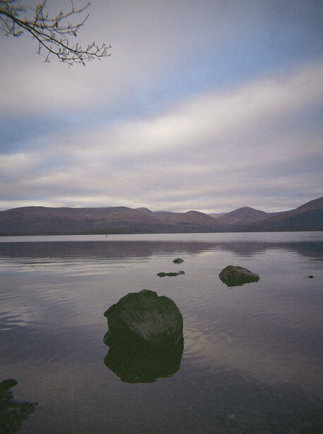 Moss-covered boulders in still water with mountains and cloudy sky beyond.