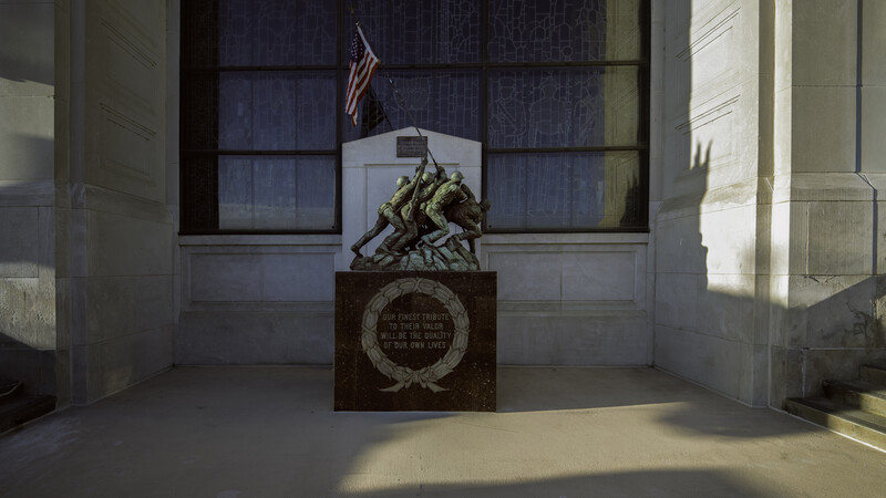 Bronze sculpture of soldiers raising a flag, positioned on a pedestal within a minimalist architectural alcove.