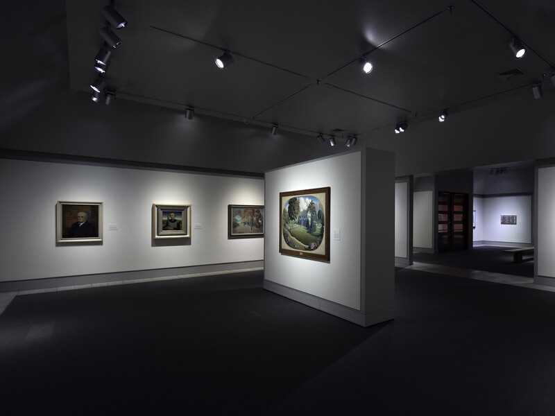 Gallery interior with framed paintings illuminated by track lighting against white walls and dark flooring.
