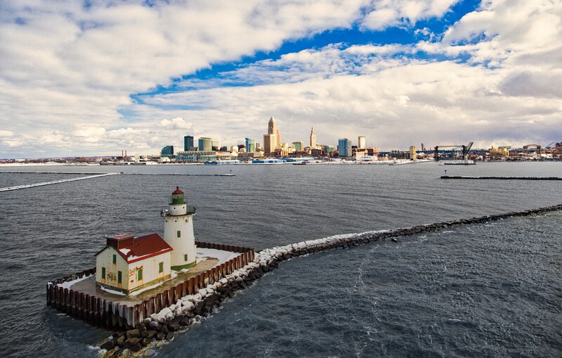 Red-roofed lighthouse on wooden pier extending into calm harbor waters with city skyline in background.