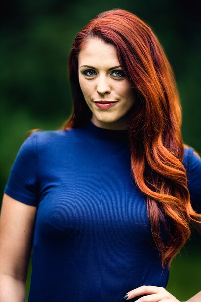 Woman with long red hair wearing a blue short-sleeved top against a blurred green background.