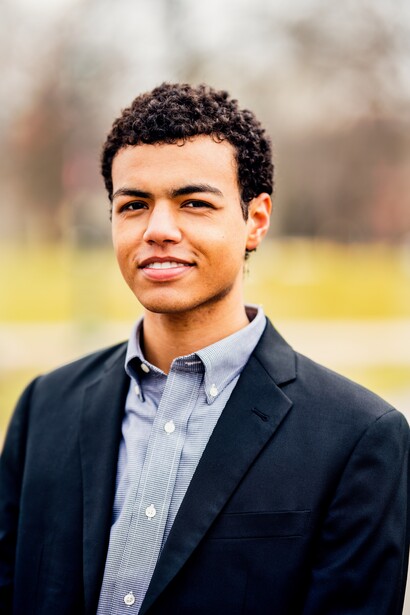 Young man in dark blazer and light blue shirt photographed outdoors with blurred background.