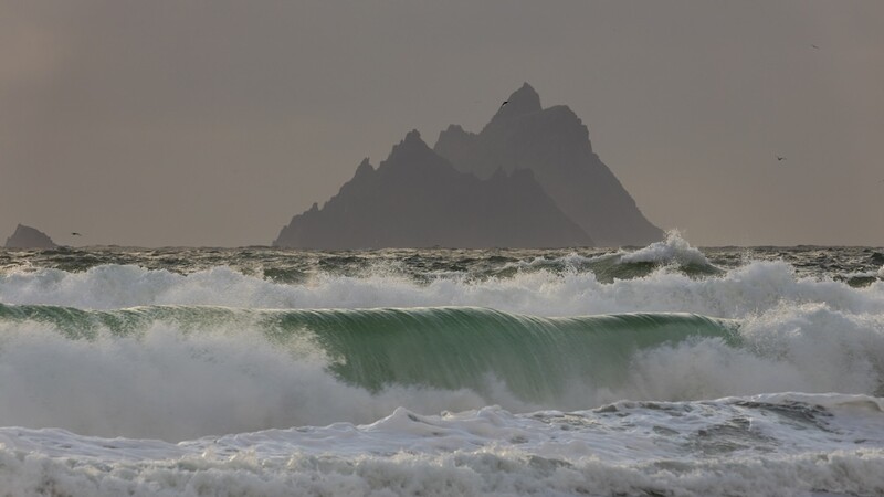 Dramatic seascape with large waves and a steep rocky mountain island silhouetted against a gray sky.