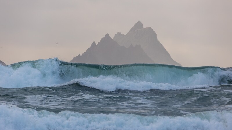 Ocean waves with a dramatic jagged mountain peak rising behind them in mist.