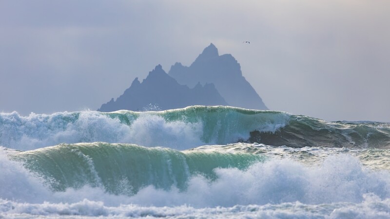 Large ocean waves with turquoise faces roll toward shore, with a jagged rocky island visible through the mist in the background.