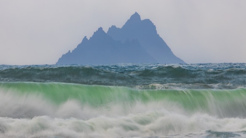 Large rocky sea stack rising from rough ocean waves with green-tinted water and white foam in foreground.