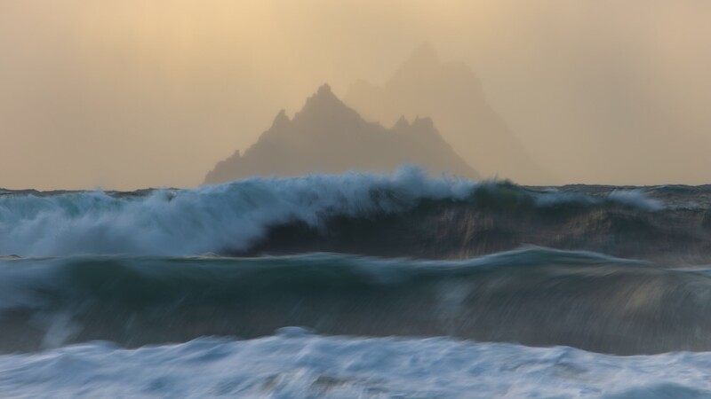 Ocean waves with white foam rolling toward shore, rocky mountain peak silhouetted in misty background.