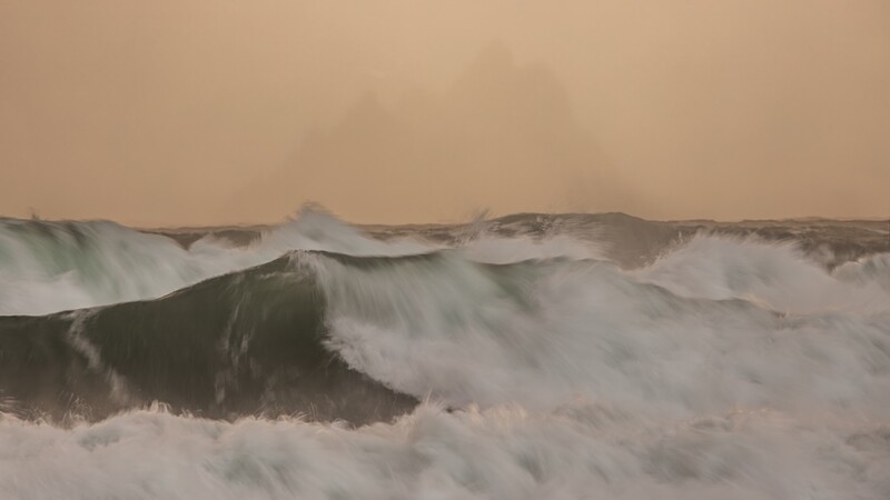 Large ocean waves with white foam rolling toward shore under a hazy, overcast sky.