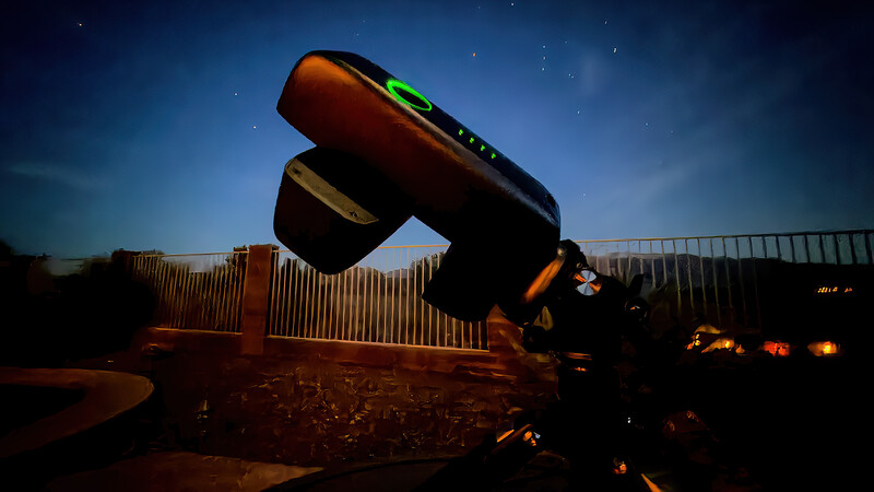 Telescope pointing upward at night sky with green laser finder visible and warm ground lighting.