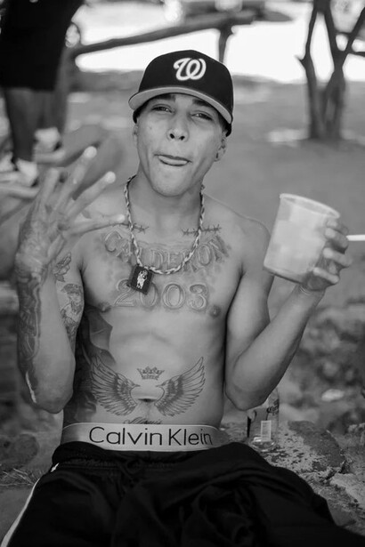 Shirtless man wearing a baseball cap and Calvin Klein underwear, holding a drink and gesturing outdoors.