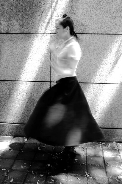 Woman in white crop top and black skirt posed against geometric wall with dramatic light and shadow patterns.