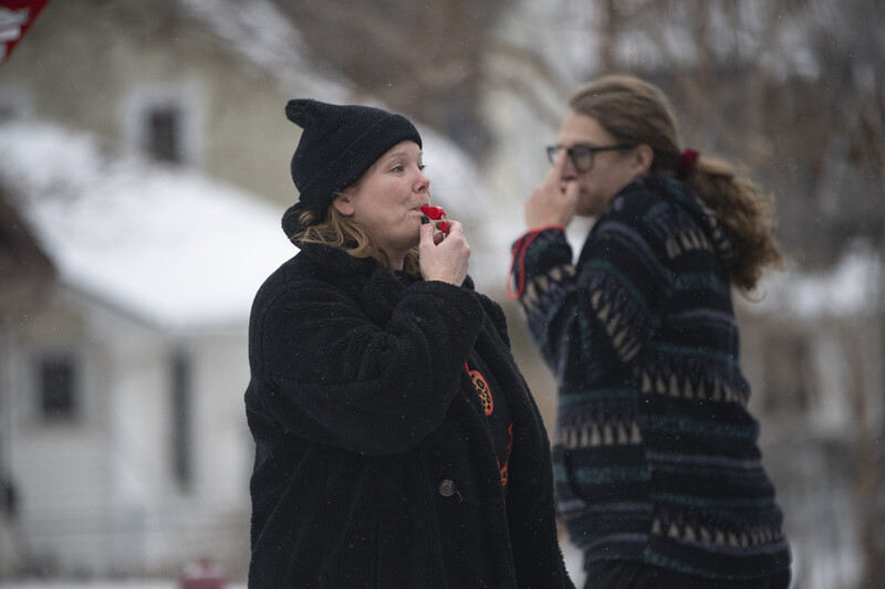 Two people in winter clothing talking outdoors with snowy residential buildings in the background.