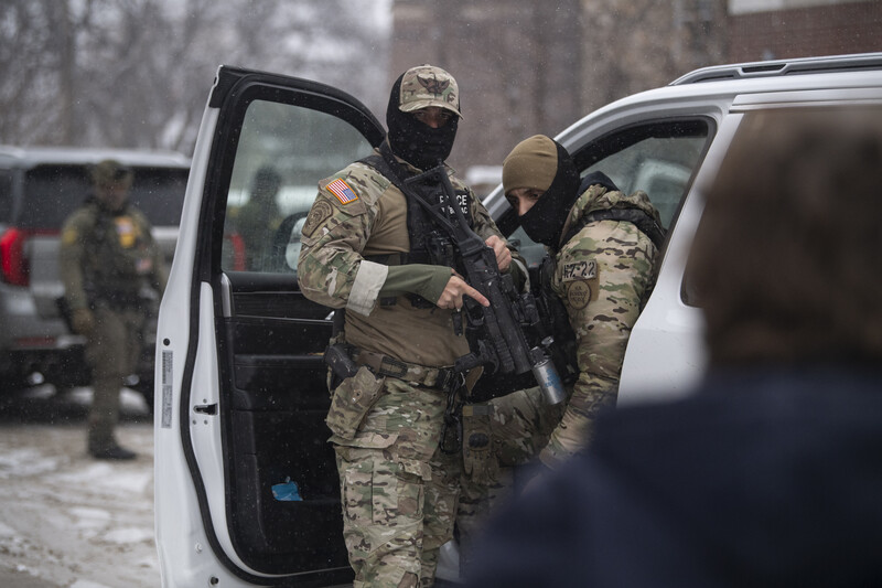 Military personnel in camouflage uniforms and tactical gear escorting a detainee from a white van.