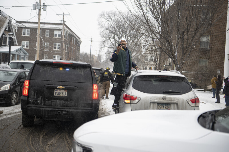 Person standing on snowy street between parked vehicles during daytime in urban neighborhood.