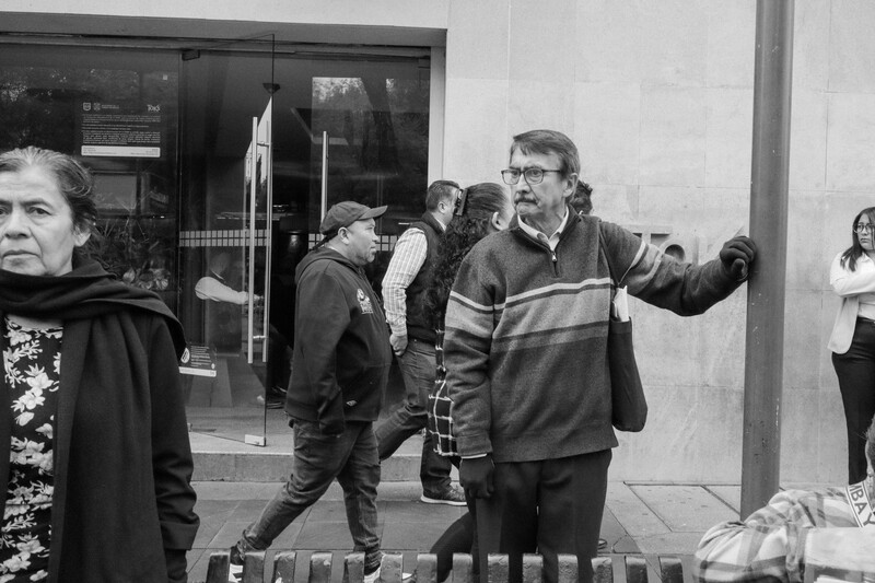 Black and white photograph of a man gesturing while speaking outdoors on a street.