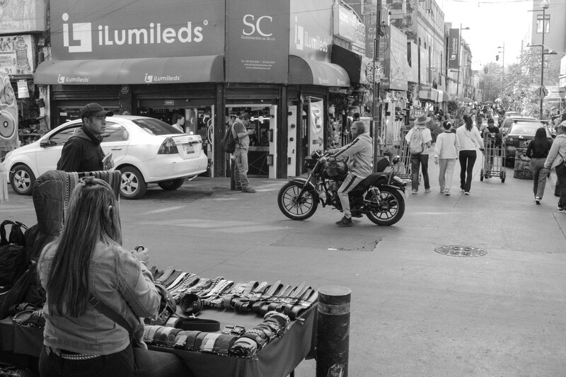 Street vendor with merchandise displayed on ground while motorcycle rider passes through busy urban sidewalk.
