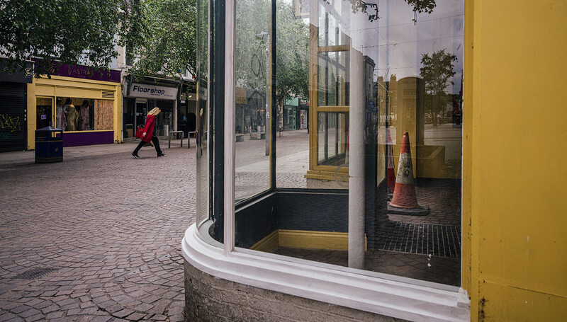Urban storefront with yellow and purple facade, curved glass window reflecting street scene and pedestrians.