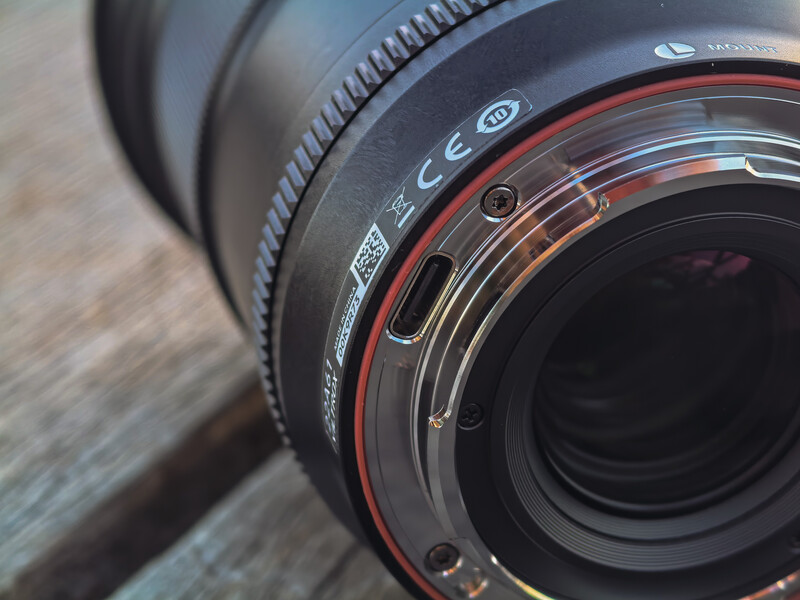 Close-up detail of a camera lens barrel showing focus ring, depth-of-field markings, and red rubber focusing ring against wooden surface.