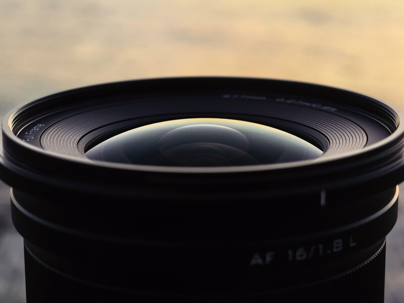 Close-up of a wide-angle camera lens with visible aperture blades and focusing ring against a blurred sky background.