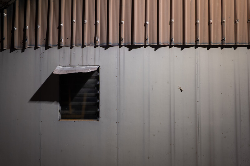 Open metal louver vent on corrugated metal wall with strong directional shadow.