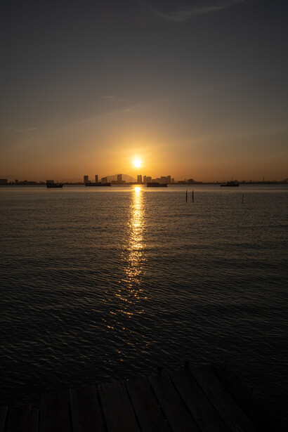 Golden sun reflecting on calm water at sunset with distant city skyline silhouetted against the horizon.