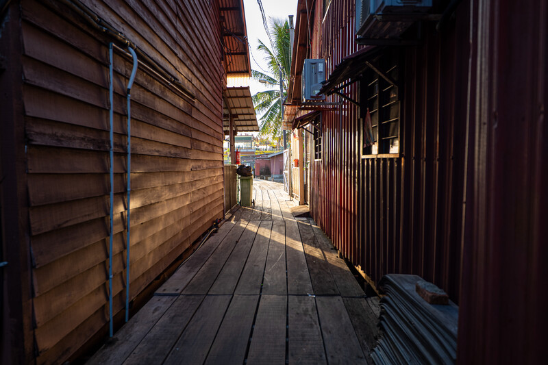 Narrow wooden boardwalk lined with weathered brown buildings in strong sunlight and shadow.