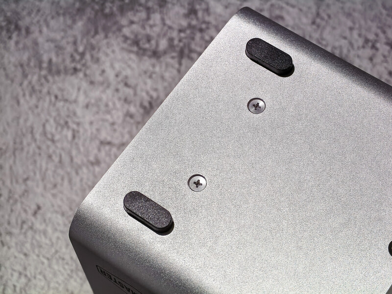 Close-up of a silver metallic device showing mounting holes and rubber feet against a textured gray surface.