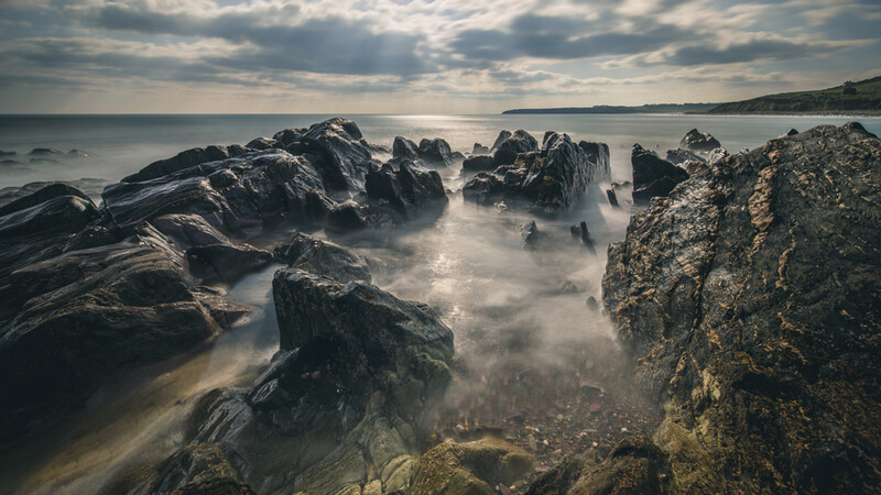 Rocky coastline with misty water flowing between dark jagged rocks under cloudy sky.