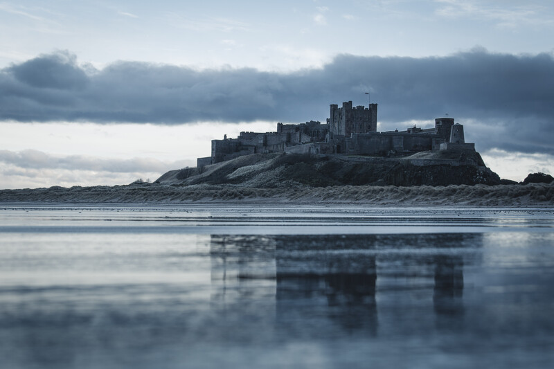 Medieval castle silhouetted on rocky island reflecting in calm tidal flats beneath stormy sky.
