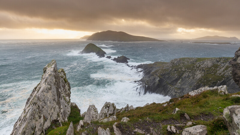 Rocky coastal headland with distant island and stormy sky over turbulent ocean.