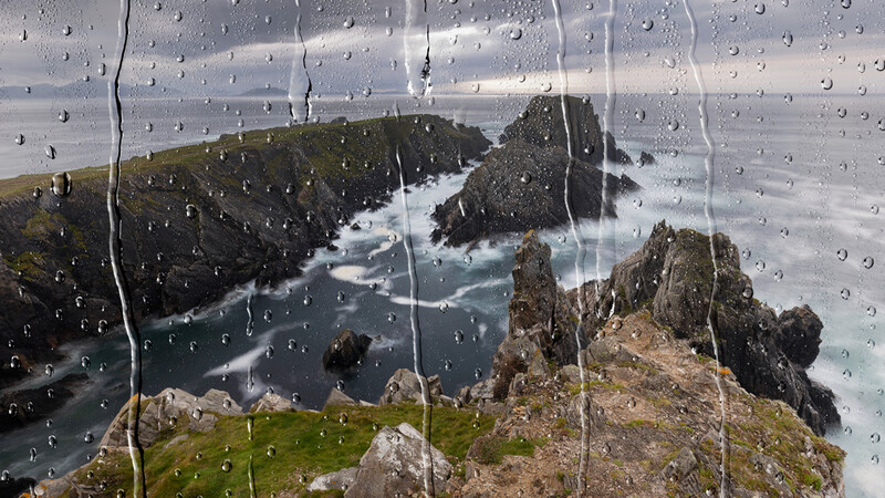 Dramatic coastal landscape with rocky cliffs and turquoise waters viewed through a rain-covered window.
