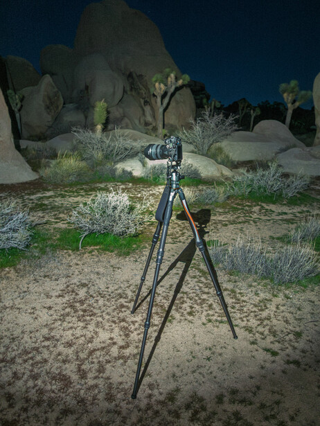 Tripod-mounted camera positioned on sandy desert ground with large boulders and sparse vegetation at dusk.
