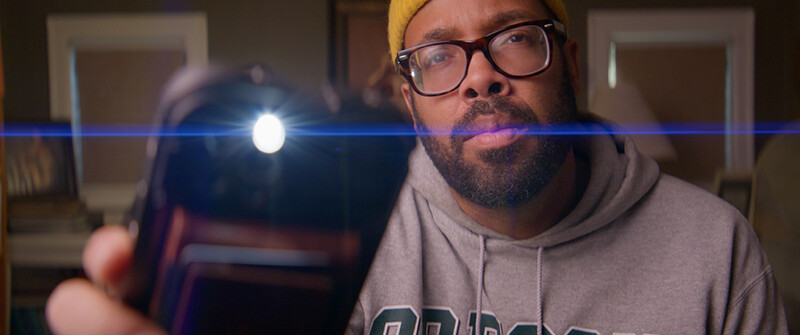 Man wearing glasses and yellow beanie holds smartphone with bright light beam crossing the frame.