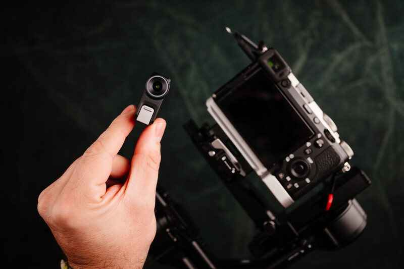 Hand holding a detached camera lens next to a mirrorless camera body on a dark green surface.
