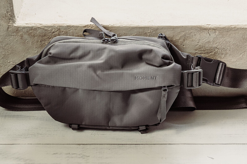 Gray camera bag with shoulder straps displayed against a white wooden wall and concrete surface.